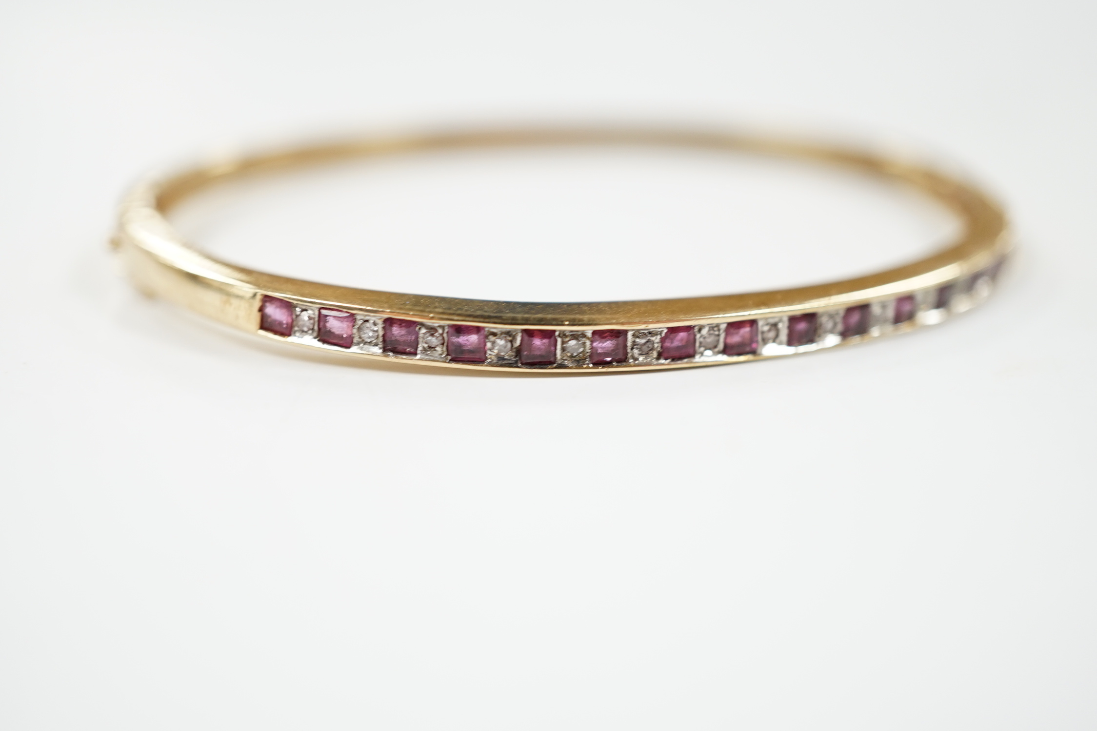 A yellow metal, ruby and diamond chip channel set hinged bangle, interior diameter 57mm, gross weight 10.8 grams.